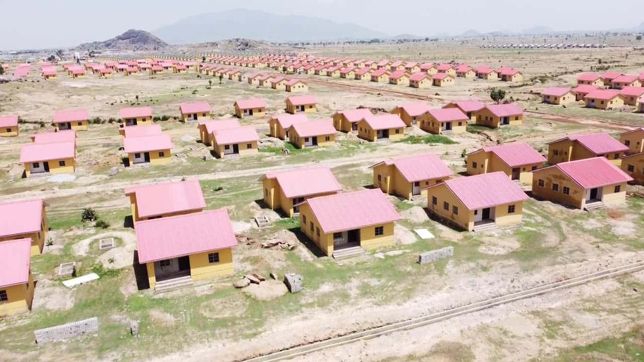 Drone view over a large housing project: identical single-story houses with pink roofs spread across open, partly cleared land.