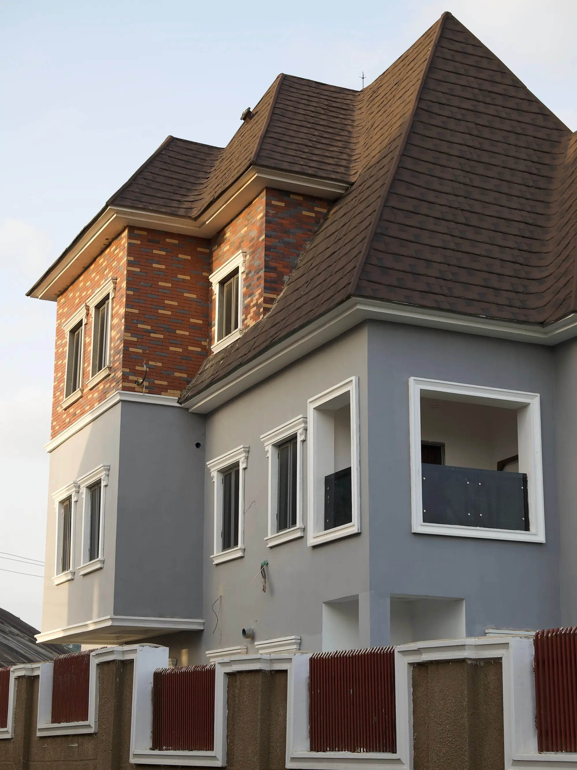 A two-story house with a brown pitched roof, a brick corner and a balcony, behind a boundary wall with red panels, under a pale sky