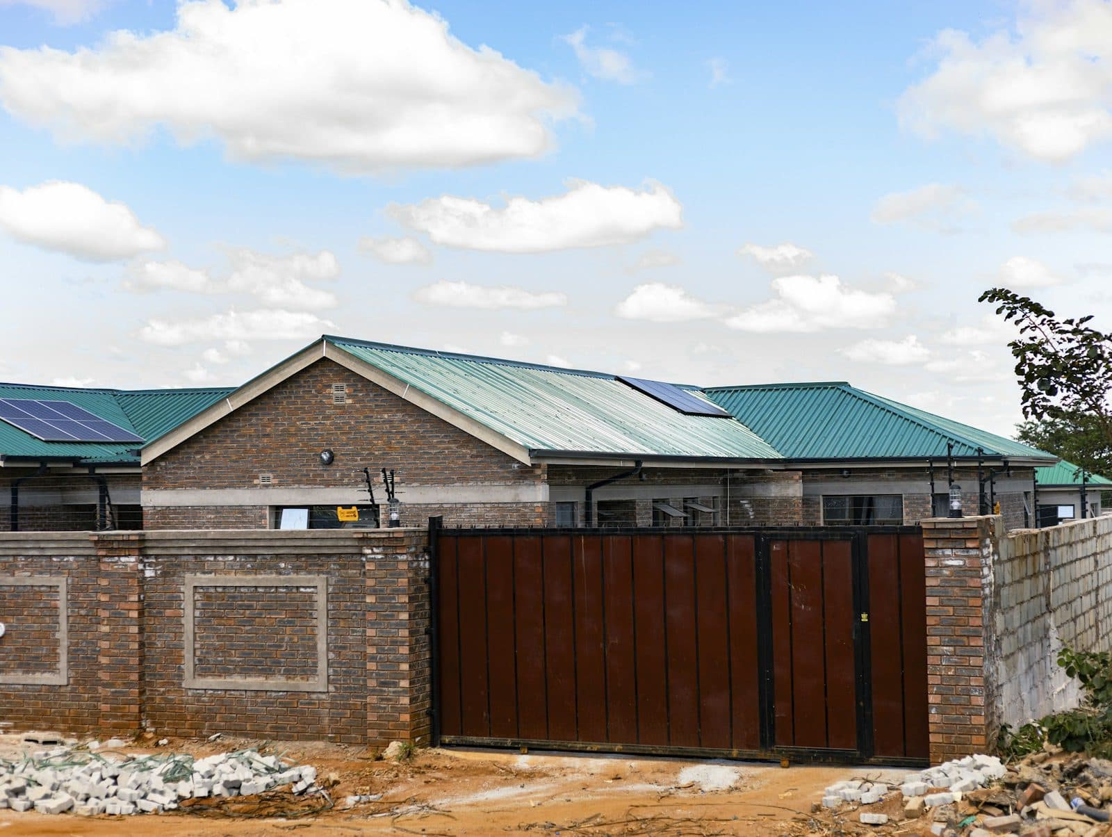 A brick boundary wall with a brown steel gate, a finished brick house with a green metal roof and solar panels behind it