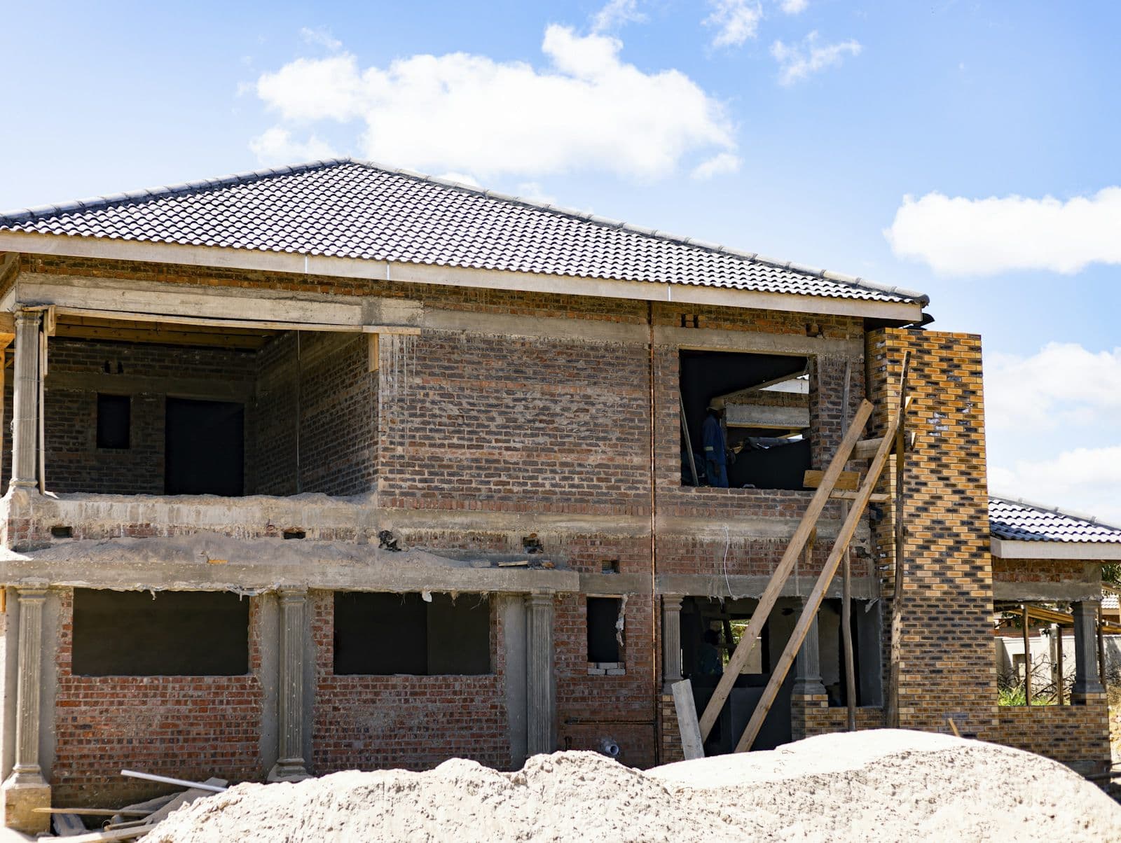 A two-storey brick house under construction with its tiled roof on, empty window openings, a ladder against the wall and heaps of sand in front