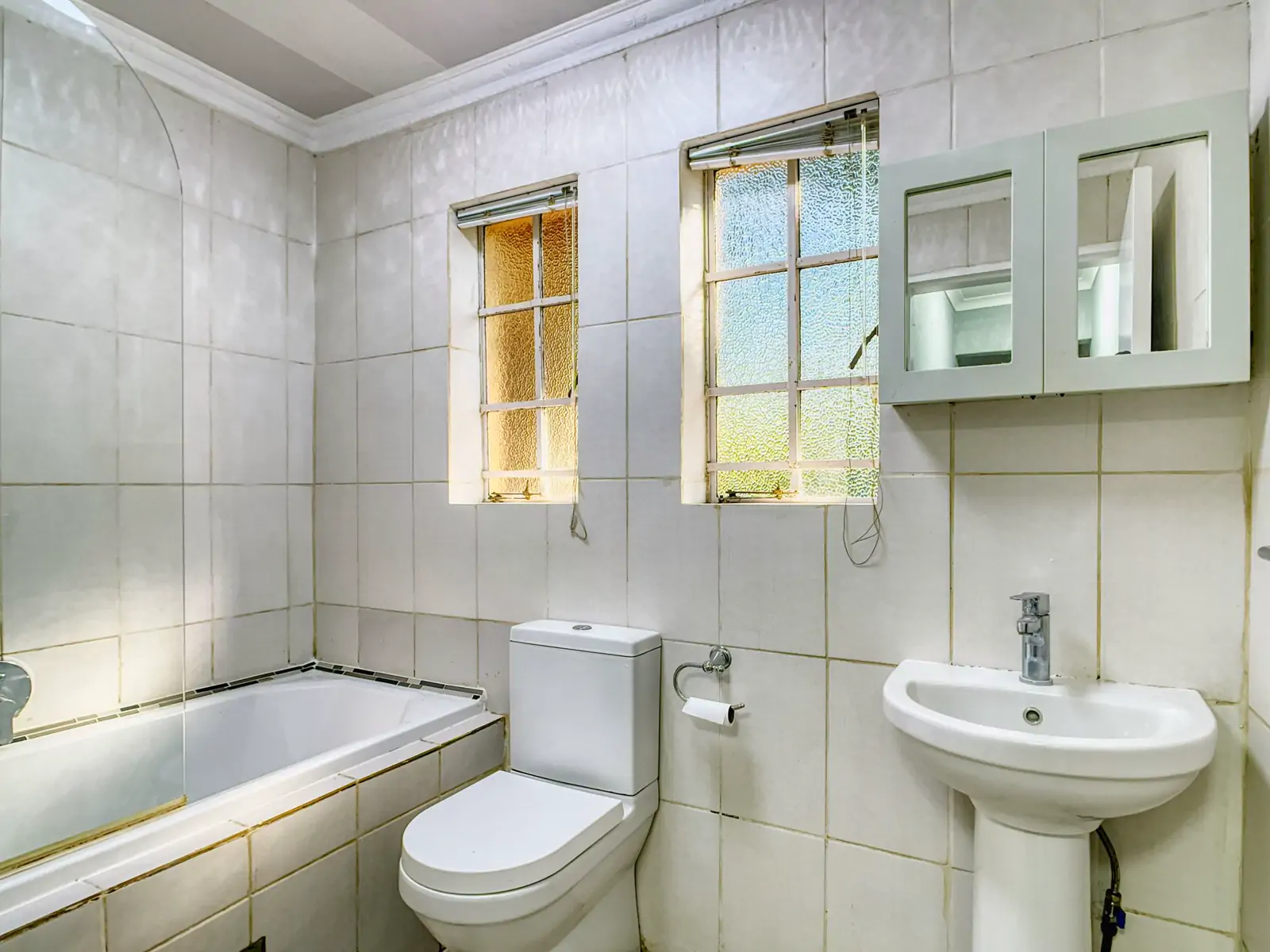 Bathroom 1: wide photo from the doorway. A white tiled bathroom with a bath, a toilet, a basin, two windows and a wall cabinet (sample photo)