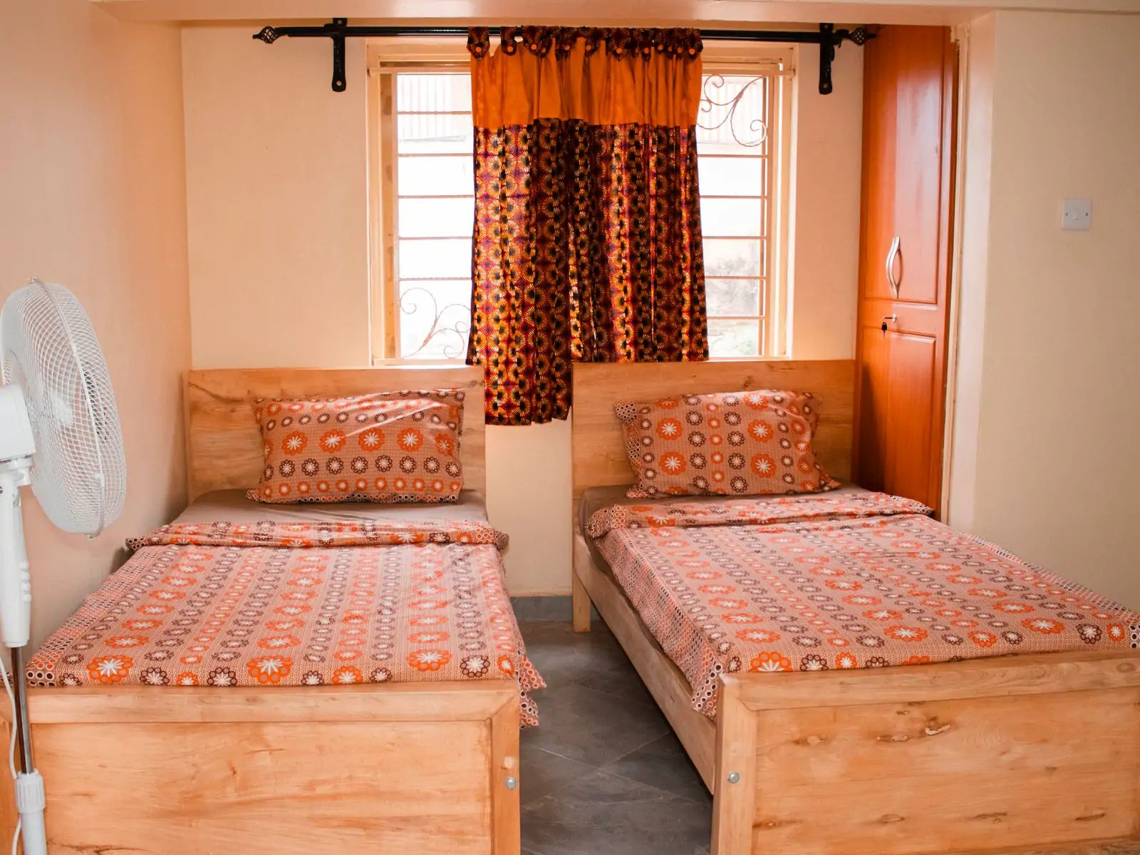 Bedroom 2: wide photo from the doorway. Two single beds with patterned covers under a window with print curtains, and a standing fan (sample photo)