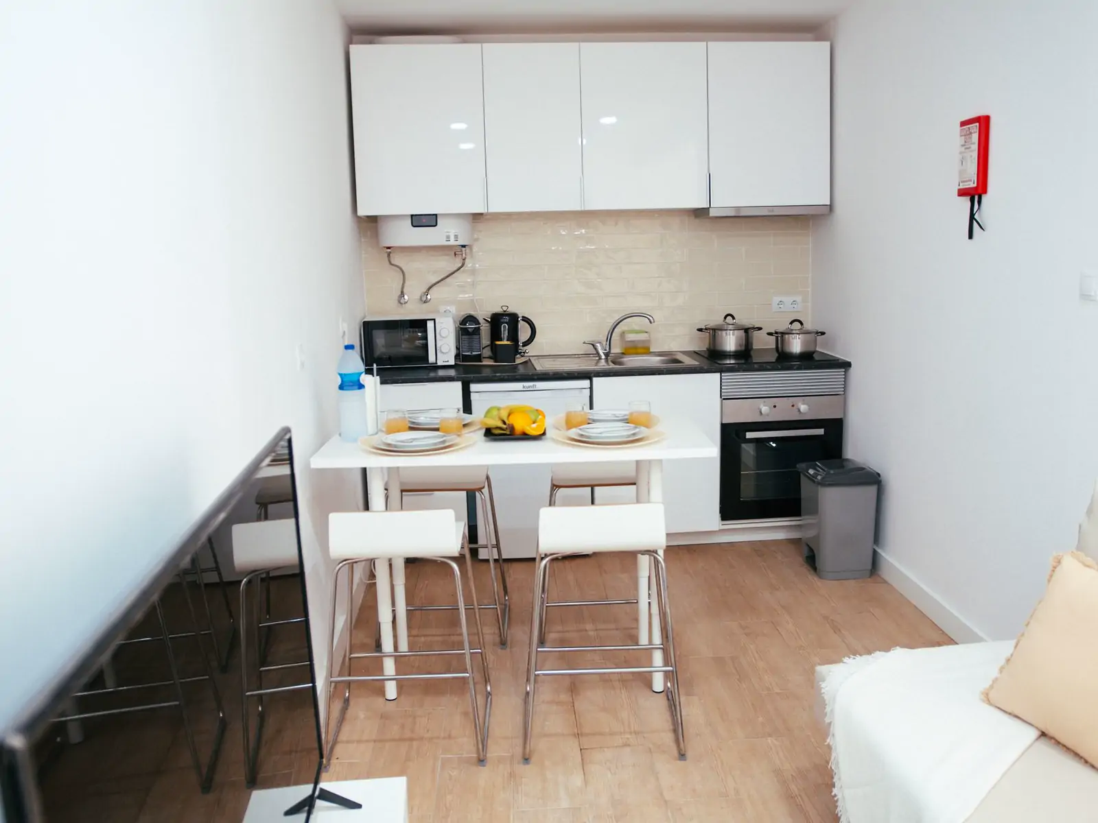 Kitchen: wide photo from the doorway. A small kitchen: white wall cupboards, a counter with a kettle and pots, an oven and a breakfast counter with two stools (sample photo)