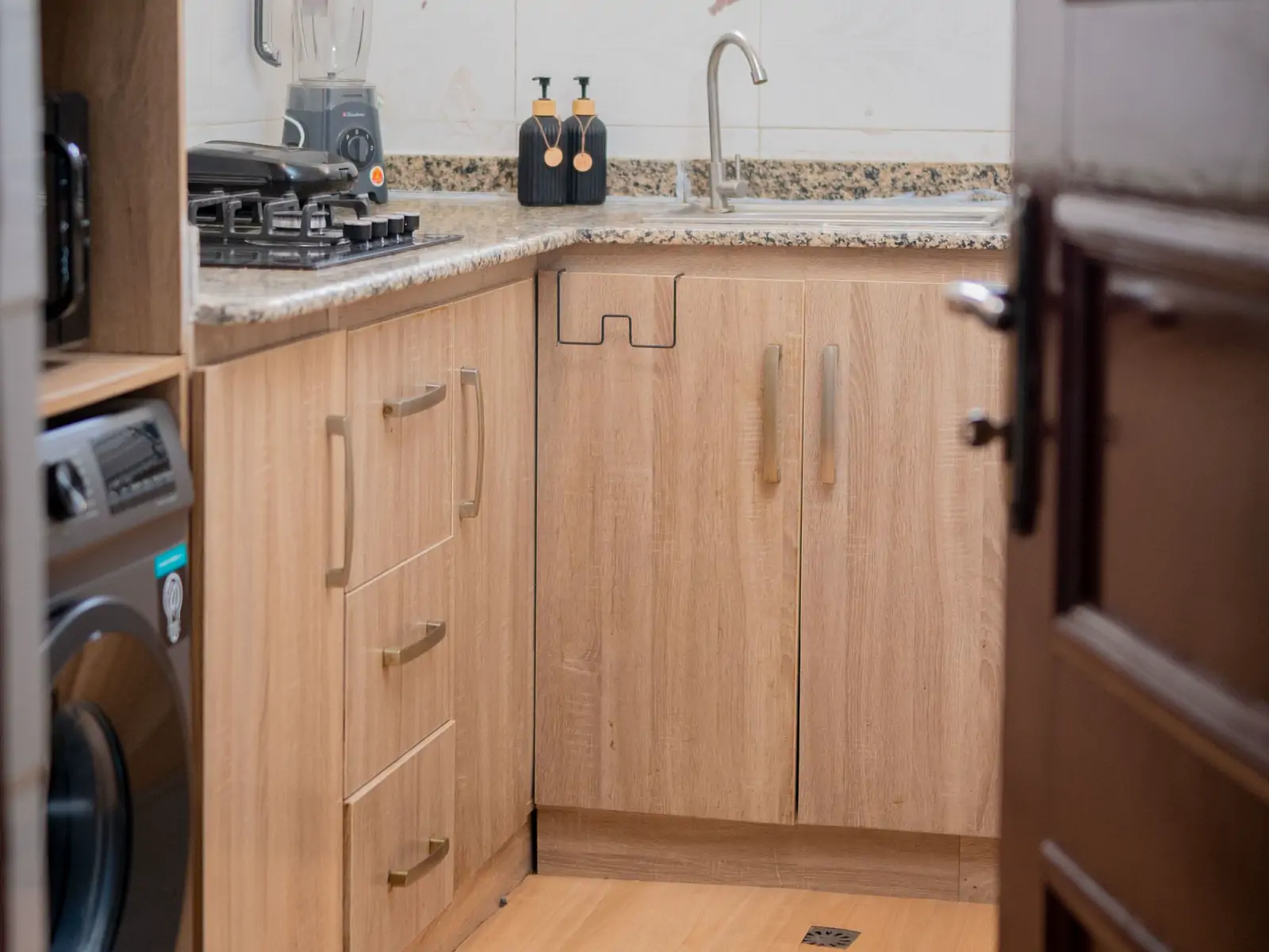 Kitchen: wide photo from the far corner. Wooden kitchen cabinets, a granite counter with a gas hob and a sink, and a washing machine (sample photo)
