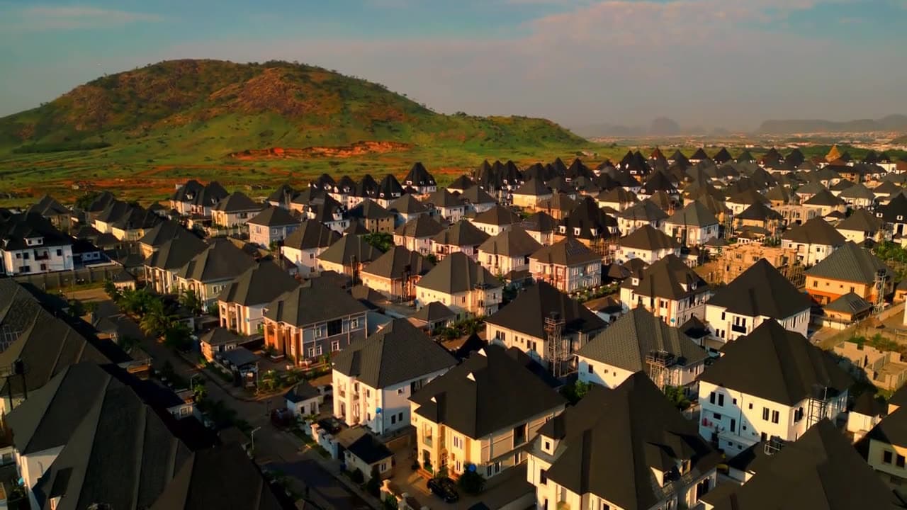Drone view over a new housing estate in Abuja in the evening sun: rows of white houses with dark roofs, a green hill behind.