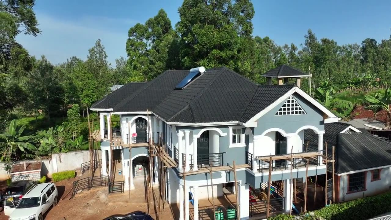 Drone view circling a large two-story house nearing completion: a dark tiled roof with a solar water heater, gray and white walls, timber scaffolding along the balconies, cars in the red-soil yard and trees all round.