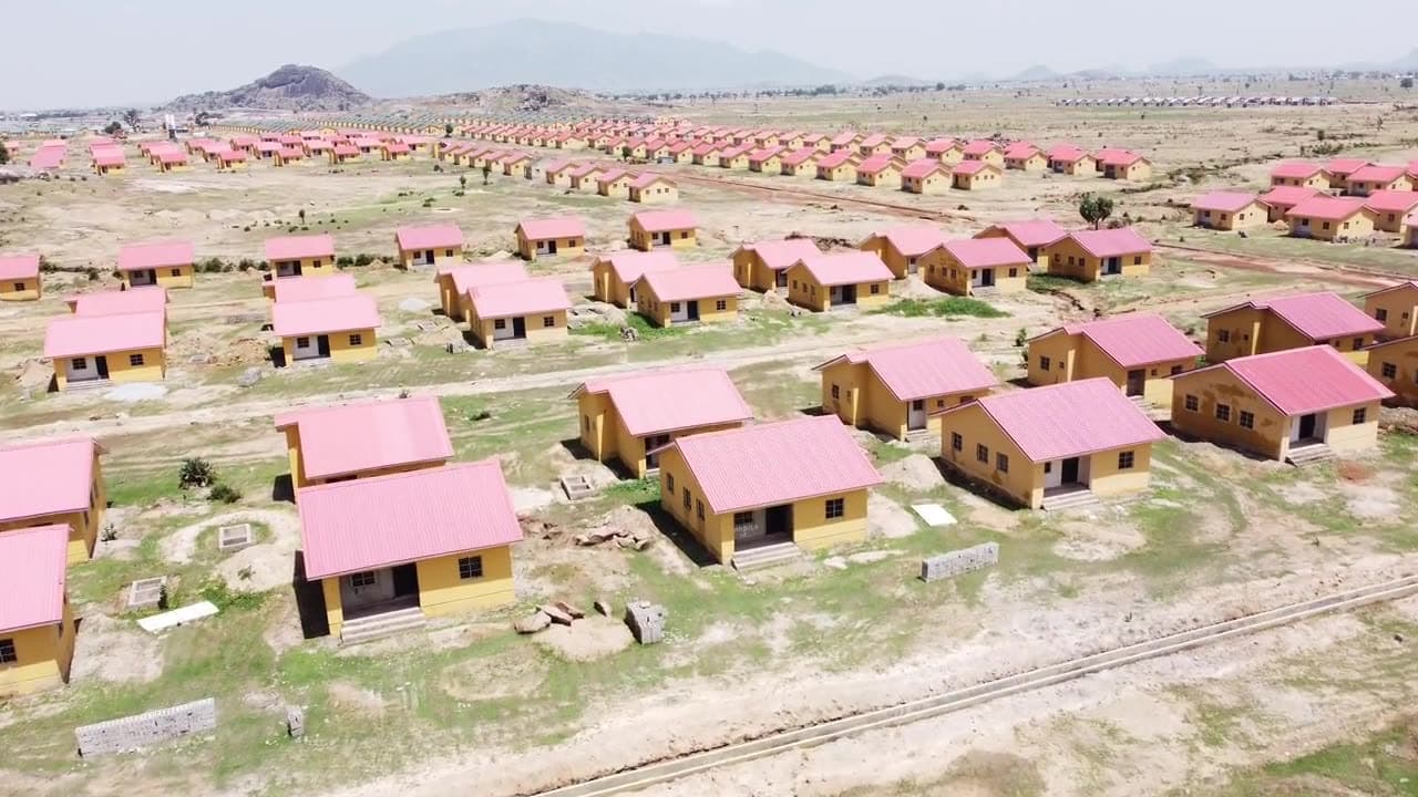 Drone view over a large housing project: identical single-storey houses with pink roofs spread across open, partly cleared land.