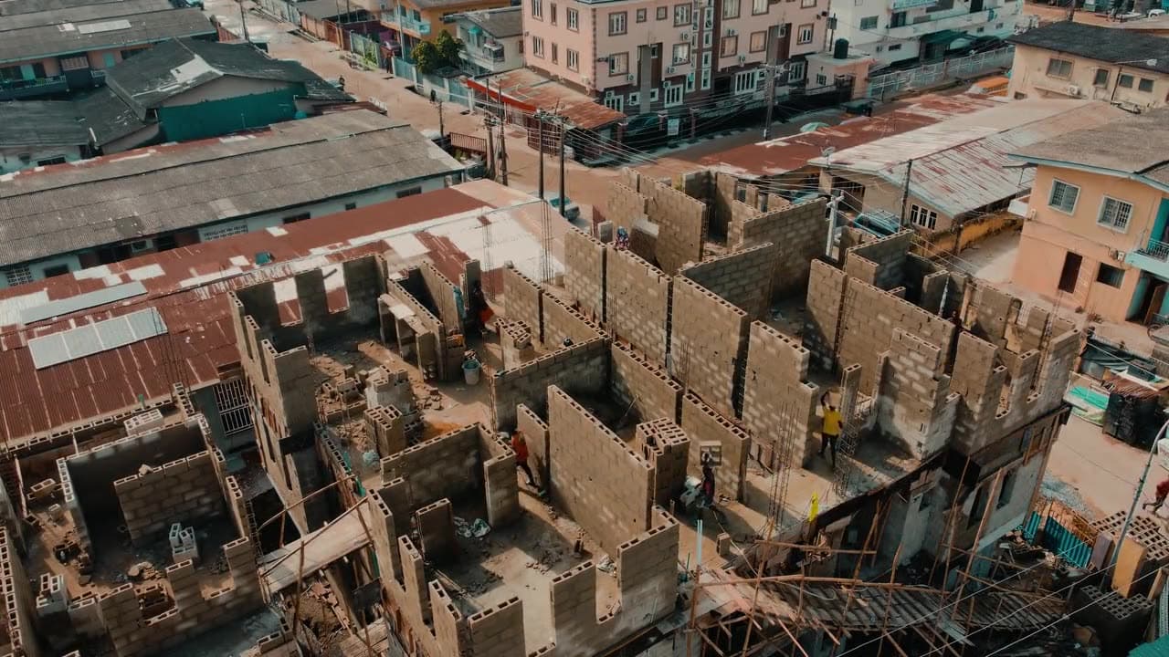 Slow drone view over a building site in Lagos: the new block walls of a many-roomed building rising between older houses with metal roofs.