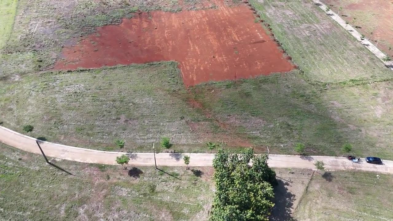 Drone view over empty building plots beside a new estate road: one cleared to red soil, the others under grass, with young trees along the road.